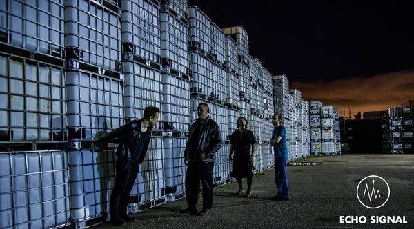 Adam, John, Phaedrus, and Charles (Echo Signal in 2016) spaced along a wall of stacked white industrial containers at night, a band of orange sky at the horizon.