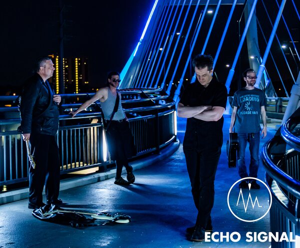 John, Phaedrus, Adam, and Charles (Echo Signal in 2016) spread along the Lowry Avenue bridge walkway at night, leaning on blue-lit railings with the lights of the Minneapolis skyline behind.